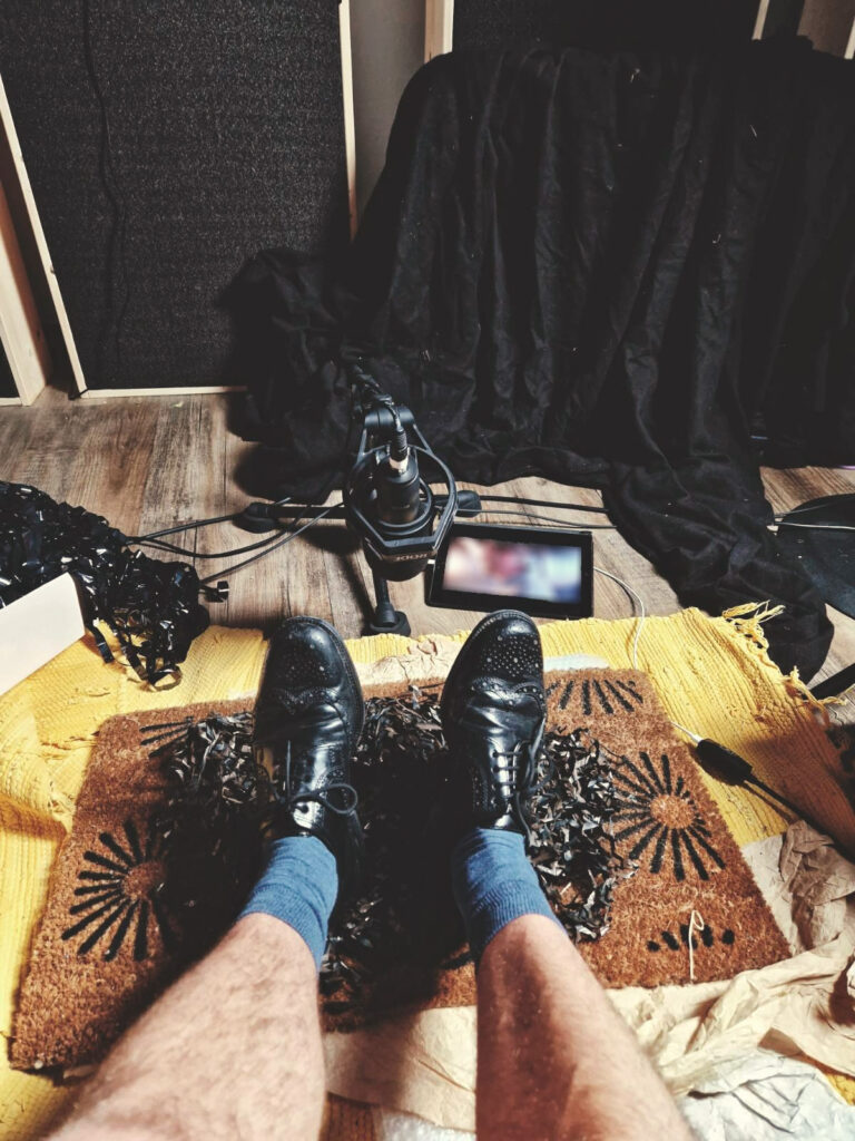 View from above: a person wearing black shoes and blue socks sits on a yellow rug decorated with a brown pattern. Theres a microphone and a tablet on a stand in front of them, surrounded by black drapery and equipment.